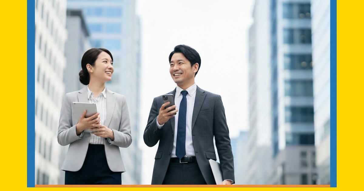 高層ビルを背景にタブレット端末やスマートフォンを携えて笑顔で話すビジネスパーソン